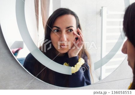 An attractive white Caucasian young woman looking in the bathroom mirror touching her wrinkles around her eyes An attractive white Caucasian young woman looking in the bathroom mirror touching her wrinkles around her eyes 136058553