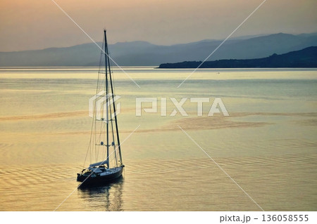 A single yacht sailing on calm waters in the early morning sunrise with distant mountains and hills in the distance A single yacht sailing on calm waters in the early morning sunrise with distant mountains and hills in the distance 136058555