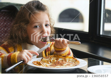 A young white Caucasian girl eating a breakfast pancake with a knife and fork 136058557