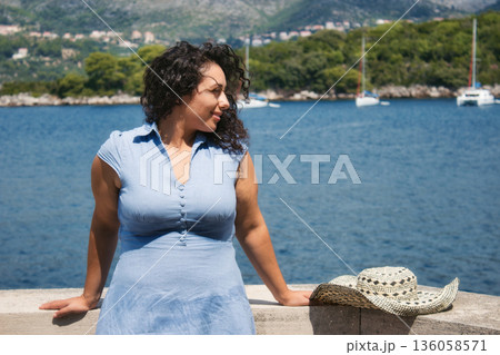 A beautiful white Caucasian woman with curly black hair wearing a blue dress, on vacation at the beach, with water and yachts visible in the background A beautiful white Caucasian woman with curly black hair wearing a blue dress, on vacation at the beach, with water and yachts visible in the background 136058571