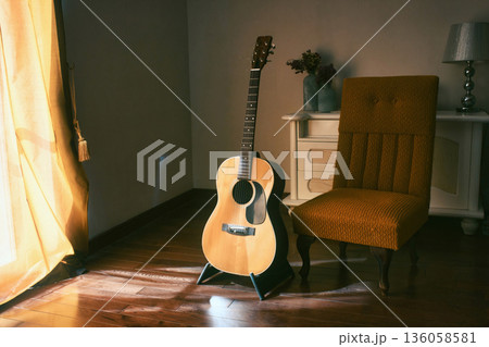An acoustic Spanish guitar on a stand next to a chair in the moody shadows of a dark room with bright light coming in from behind a curtain 136058581