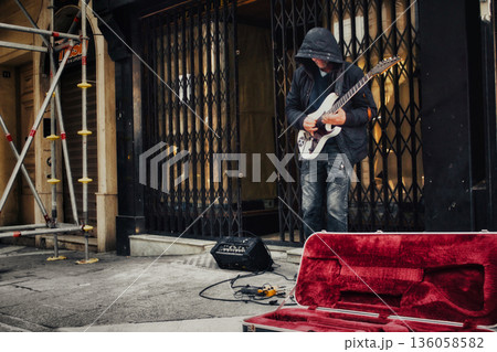 An unrecognizable hooded busker in a pedestrian street in a city center playing an electric blues guitar with an amplifier and effects pedals An unrecognizable hooded busker in a pedestrian street in a city center playing an electric blues guitar with an amplifier and effects pedals 136058582