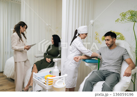 Wide shot of medical professional in uniform and gloves prepares male patient arm for blood draw while colleague having consultation with client in background, depicting routine diagnostic testing. 136059196