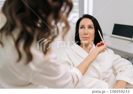 Back view of experienced beauty technician consulting with woman seated comfortably in chair, using pencil to outline features for upcoming cosmetic enhancement at contemporary beauty center. 136059233