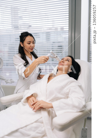 Vertical shot of cosmetic professional doctor performing rejuvenating ultrasound lifting facial treatment on adult woman face at modern aesthetic clinic, focusing on antiaging and skin contouring. Vertical shot of cosmetic professional doctor performing rejuvenating ultrasound lifting facial treatment on adult woman face at modern aesthetic clinic, focusing on antiaging and skin contouring. 136059257