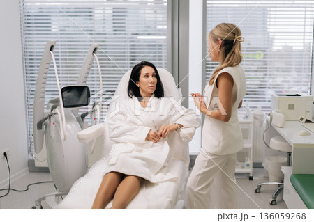 Cosmetologist consulting female client about skin rejuvenation treatment using advanced ultrasound device in modern dermatological clinic, discussing options for beauty care. Cosmetologist consulting female client about skin rejuvenation treatment using advanced ultrasound device in modern dermatological clinic, discussing options for beauty care. 136059268
