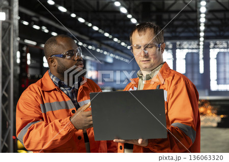 Diverse operators use laptop to coordinate production activity on the factory floor, highlighting manpower and the industrial environment for heavy manufacture and logistic tasks. Diverse operators use laptop to coordinate production activity on the factory floor, highlighting manpower and the industrial environment for heavy manufacture and logistic tasks. 136063320