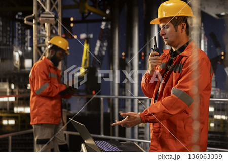 Skilled offshore engineer talking through walkie talkie at night for drilling rig systems monitoring activity and promoting petrol control and extraction practices. Oil pumping. 136063339