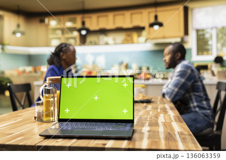 Chroma key display on laptop next to laughing couple enjoying morning routine together at the kitchen table. Man and woman drinking coffee, symbolizing communication and household relaxation. Chroma key display on laptop next to laughing couple enjoying morning routine together at the kitchen table. Man and woman drinking coffee, symbolizing communication and household relaxation. 136063359