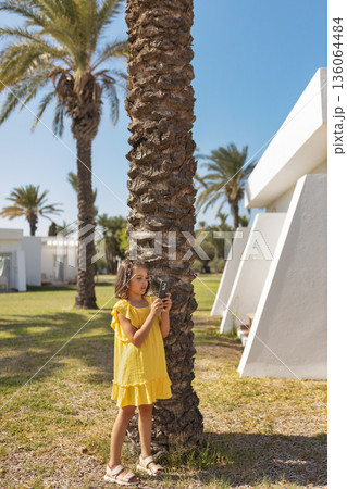 Happy cheerful girl 7 years old in yellow dress looks at phone on lawn with palm trees in summer day, life style Happy cheerful girl 7 years old in yellow dress looks at phone on lawn with palm trees in summer day, life style 136064484
