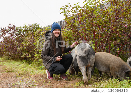 Russian woman farmer smiling at the camera while petting a mini pig on a farm. Happy rural lifestyle and connection with animals outdoors. 136064816