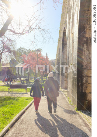 Old people walking along path beside historic aqueduct in sunny city park backlit sun and long shadows leisure scene travel lifestyle concept. High quality photo 136065061