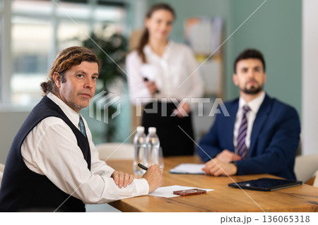Business partners sitting at table and chatting, colleague with remote control telling at background 136065318