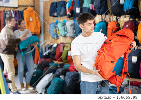 Guy chooses and buys tourist backpack for summer hike in sports equipment store 136065599