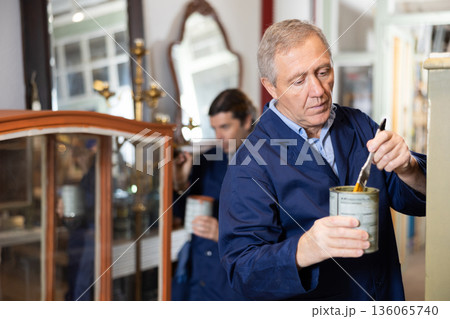 Skilled old furniture repair specialist in uniform restoring and painting cupboard furniture during workday in workshop 136065740