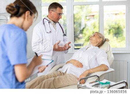Doctor with female assistant talking to elderly woman lying on examination couch 136065856
