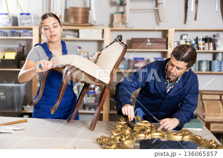 Male restorer treats wooden frame of vintage mirror with protective varnish. While woman restorer restores upholstery of vintage chair Male restorer treats wooden frame of vintage mirror with protective varnish. While woman restorer restores upholstery of vintage chair 136065857
