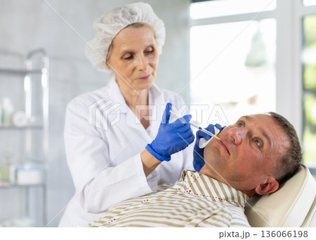 Female cosmetologist performing filler injections into lips of man 136066198
