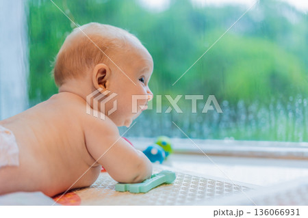Little baby looking through a window while rain falls outside, quiet childhood moment filled with curiosity and calm. Cozy home atmosphere, early development, emotions, and reflection concept 136066931