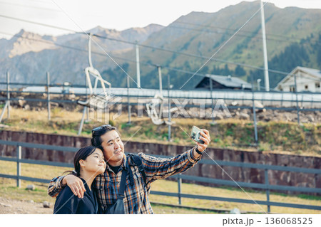 young Asian couple stands together in a mountainous area. They smile while take a selfie near a cable car station, enjoying their vacation in nature. 136068525