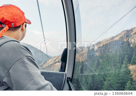 young Asian boy with short black hair wearing a red cap looks out of a cable car at mountains and trees during vacation. 136068604