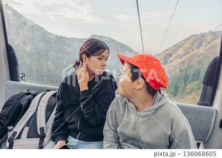 Two children ride a cable car in the mountains. A young Asian boy with a red cap and a young Asian girl smile together. Scenic mountain view in the background Two children ride a cable car in the mountains. A young Asian boy with a red cap and a young Asian girl smile together. Scenic mountain view in the background 136068605