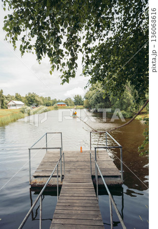 A wooden pier extends into a serene river surrounded by lush greenery. Calm waters reflect the sky, creating a peaceful atmosphere. 136068616