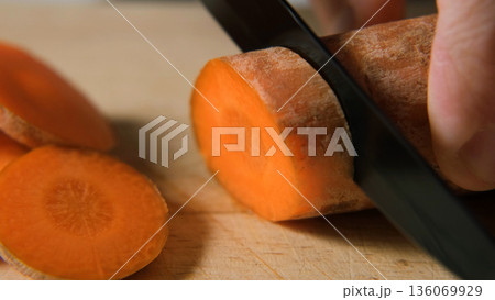 Macro of slicing carrot with knife on wooden surface, showing texture and color of fresh vegetable. Healthy cooking concept Macro of slicing carrot with knife on wooden surface, showing texture and color of fresh vegetable. Healthy cooking concept 136069929