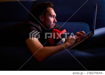 Casual night working from comfy sofa, Quiet evening spent on freelance project in cozy living room, Man enjoying late night work at home with relaxed posture and soft glow 136070644