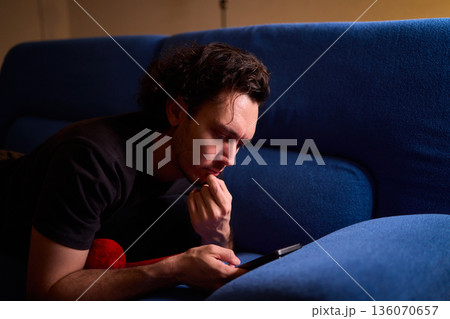 Chilled man on sofa, Casual individual reclining on sofa engrossed with mobile device, Unwinding man sits comfortably on sofa focused on his smartphone amid cozy modern decor 136070657