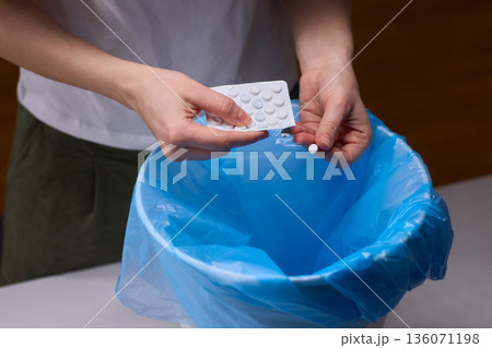 Hands discarding expired pills into bin, closeup of blister pack over blue plastic liner, tabletop setting, focused on safe disposal, mindful household routine, low warm lighting Hands discarding expired pills into bin, closeup of blister pack over blue plastic liner, tabletop setting, focused on safe disposal, mindful household routine, low warm lighting 136071198