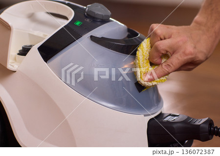 Hand cleaning steam cleaner reservoir with yellow sponge, green indicator light glowing, closeup of plastic tank and handle, focus on routine maintenance and hygiene in modern home 136072387
