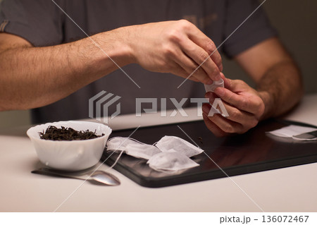 Artisan tea bag filling process, Careful and detailed artisanal approach to tea packaging, Meticulous handpicked tea bags with loose leaves and scattered empty sachets on tray 136072467