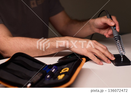 Hands applying solder paste to board, closeup of repair workspace with soldering iron, tool pouch, black device, soft warm lighting, focused precision work, hobbyist repair scene Hands applying solder paste to board, closeup of repair workspace with soldering iron, tool pouch, black device, soft warm lighting, focused precision work, hobbyist repair scene 136072472