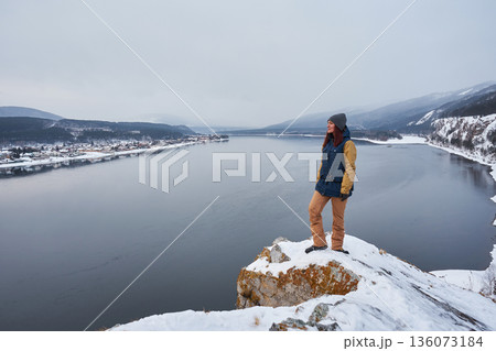 Woman tourist on a snowy hilltop overlooking the Yenisei River and a winter village. Winter adventure travel and exploration concept. 136073184