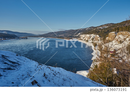 Blue water of the Yenisei River flowing through a winter landscape with snow-covered banks and distant mountains under a clear sky. 136073191