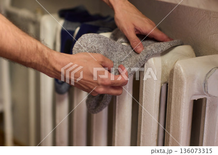 Person placing moist wool socks onto heated radiator fins, Detailed scene of hand carefully positioning damp wool socks on heated metal radiator with knit texture 136073315