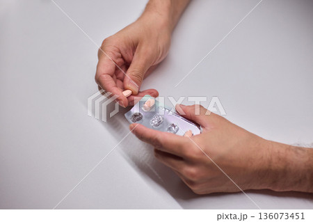 Hands extracting capsule from blister pack gently, closeup on fingertips and foil on white tabletop, quiet moment of dosing accuracy and calm preparation for intake Hands extracting capsule from blister pack gently, closeup on fingertips and foil on white tabletop, quiet moment of dosing accuracy and calm preparation for intake 136073451