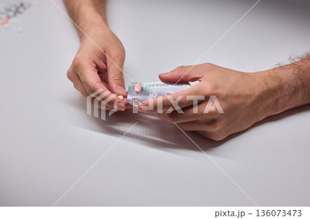 Hands extracting capsule from blister pack gently, closeup on fingertips and foil on white tabletop, quiet moment of dosing accuracy and calm preparation for intake Hands extracting capsule from blister pack gently, closeup on fingertips and foil on white tabletop, quiet moment of dosing accuracy and calm preparation for intake 136073473