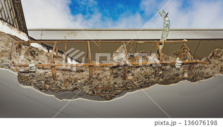 Close-up of crumbling reinforced concrete with exposed rusty rebar and aggregates under a building roof. Close-up of crumbling reinforced concrete with exposed rusty rebar and aggregates under a building roof. 136076198