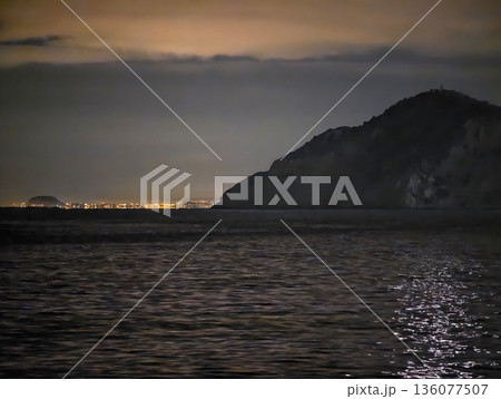 Nighttime coastal vantage showcases glowing city reflections and tranquil yet alert atmosphere Nighttime coastal vantage showcases glowing city reflections and tranquil yet alert atmosphere 136077507