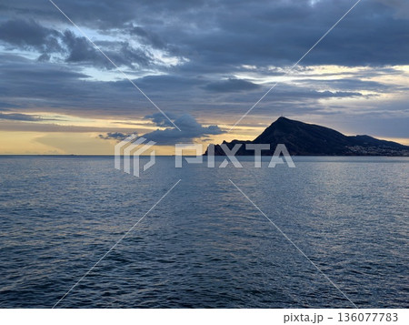 Calm sea with island silhouette at sunset, Tranquil ocean view with distant mountains and layered clouds 136077783