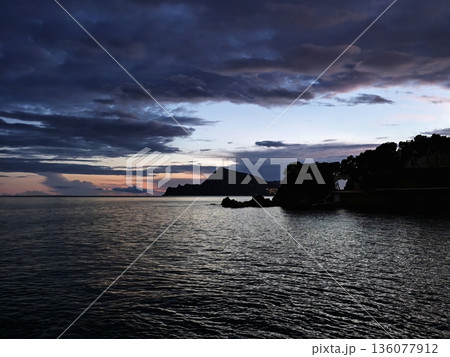 Tranquil harbor city nestled beneath mountains as evening falls and lights sparkle on water 136077912