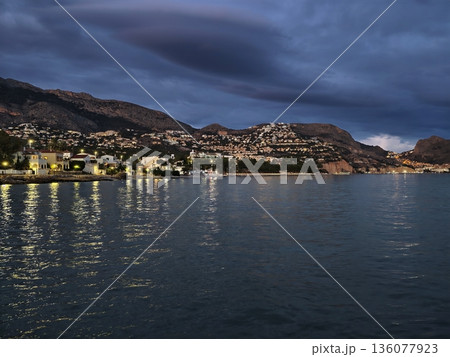Serene and undisturbed sea reflects faint glow of distant city lights over gentle rolling hills Serene and undisturbed sea reflects faint glow of distant city lights over gentle rolling hills 136077923