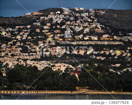Tranquil coastal mountain town illuminated by evening lights reflecting on calm water surface Tranquil coastal mountain town illuminated by evening lights reflecting on calm water surface 136077928