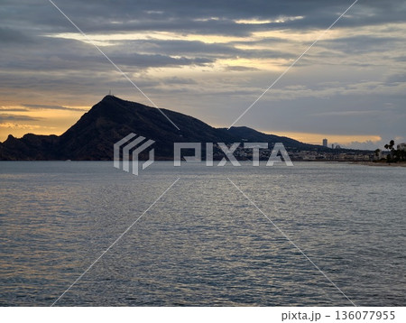 Calm island sunset scene, Quiet sunset over tranquil sea with distant village lights and clouds Calm island sunset scene, Quiet sunset over tranquil sea with distant village lights and clouds 136077955