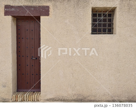 Minimalist composition highlighting tall wooden door beside barred window on muted plaster facade Minimalist composition highlighting tall wooden door beside barred window on muted plaster facade 136078035