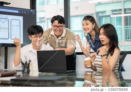 Asian man pointing and coworkers making a fist while looking at laptop on glass top table in office. Asian man pointing and coworkers making a fist while looking at laptop on glass top table in office. 136078899