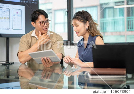 Asian glasses man holding tablet and stylus pen talking to woman coworker in office's meeting room. Asian glasses man holding tablet and stylus pen talking to woman coworker in office's meeting room. 136078904