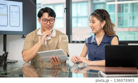Asian glasses man holding tablet and stylus pen talking to a woman coworker in office's meeting room 136078905